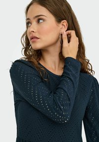 Námořnické šaty s dlouhým rukávem a texturovaným designem v chevronovém vzoru. Modelka nosí stříbrný kruhový náušník a jednou rukou se dotýká ucha.
