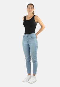 Schwarzes ärmelloses Oberteil, hellblaue hochtaillierte Jeans, weiße Sneakers. Das Model steht mit einer Hand auf der Hüfte, lässiger Schnitt und leichte Denim-Textur.