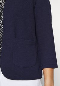 Marinblå texturerad jacka med en liten ficka fram och trekvarts ärmar, med en öppen frontdesign och en mjuk foder.