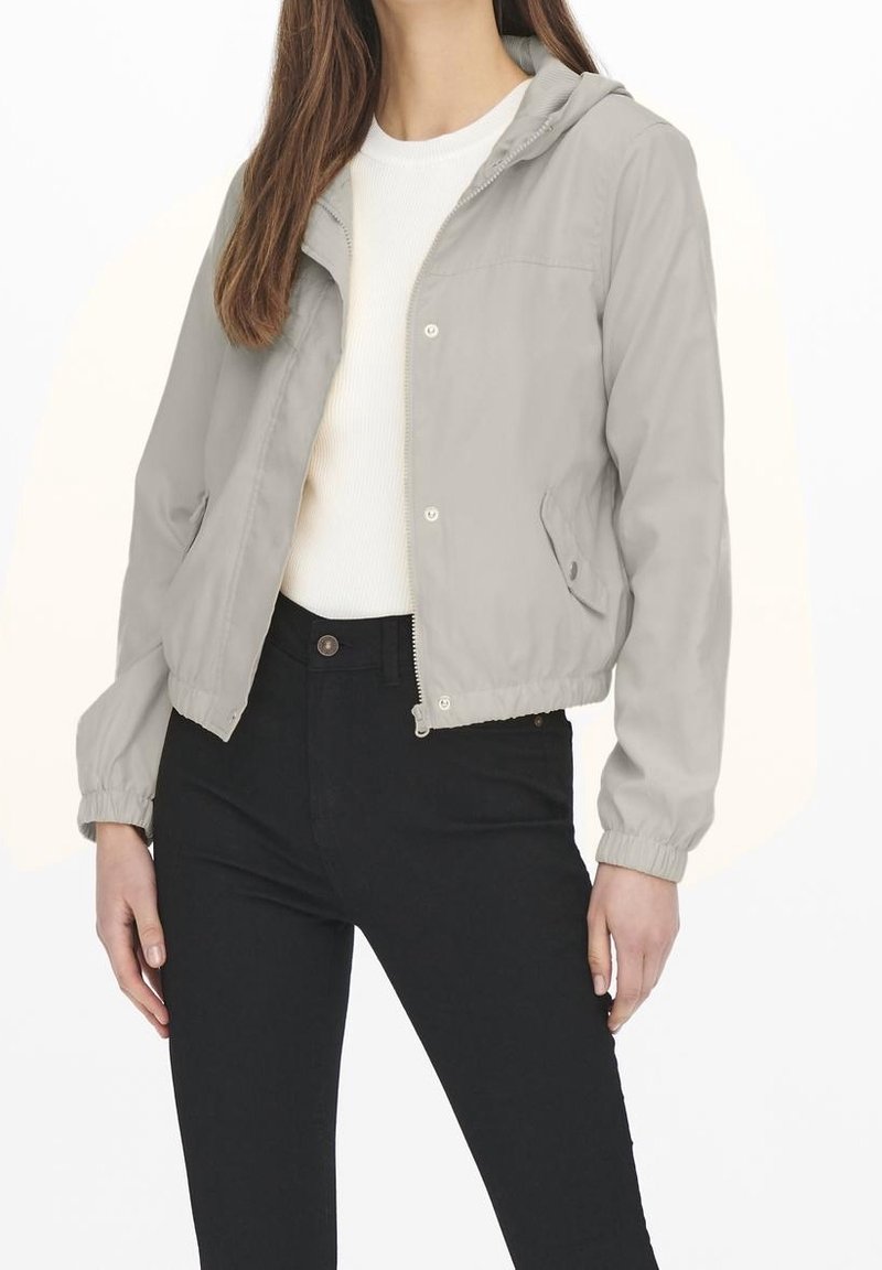 Světle šedá lehká bunda s kapucí, která má zapínání na zip v přední části, pružné manžety a boční kapsy, je kombinována s bílým topem a černými džíny.