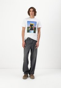 White graphic t-shirt featuring a colorful print, paired with dark denim jeans and tan shoes. Simple design, relaxed fit, and casual styling.