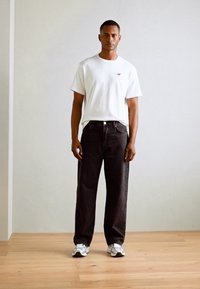 Hvid bomulds t-shirt med et lille rødt logo, parret med sorte jeans. Model bærer hvide sneakers og står på et trægulv mod en lys væg.