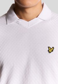 Vit polotröja med textilstruktur, featuring en v-ringning och en gul broderad fågel-logotyp på vänster bröst.