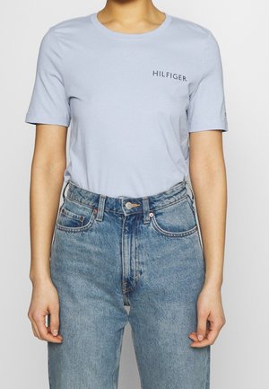 Person trägt hellblaues T-Shirt mit kurzen Ärmeln mit dem Text "HILFIGER" und hochgeschnittene blaue Jeans vor einfachem Hintergrund.