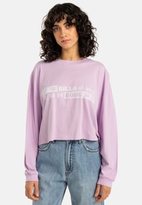 Lavender long-sleeve cropped t-shirt with "BILLA BONG" text and floral accents, paired with light blue denim jeans. Soft cotton material.