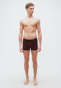 Νέος άνδρας όρθιος, ξυπόλητος, φορώντας σκούρα καφέ μποξεράκια, κοιτάζοντας ελαφρώς προς τα αριστερά του, μπροστά σε απλό λευκό φόντο.