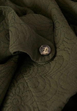 Tissu vert olive avec un motif tourbillonnant texturé, comportant un bouton en plastique clair à quatre trous. Apparence douce et drapée.