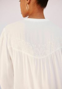 Weiße Bluse mit einem bestickten Muster am Rücken-Halsausschnitt. Weicher, fließender Stoff mit Faltendetails am unteren Saum.