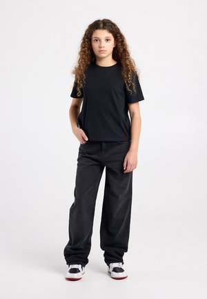 T-shirt noir à col rond, assorti à un jean noir ample et des baskets noires avec des accents blancs. Cheveux bouclés tombant en cascade.