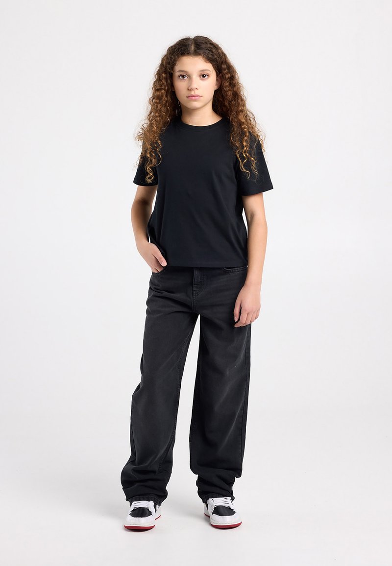 Schwarzes T-Shirt mit rundem Halsausschnitt, kombiniert mit locker sitzenden schwarzen Jeans und schwarzen Sneakers mit weißen Akzenten. Lockiges Haar, das herabfällt.