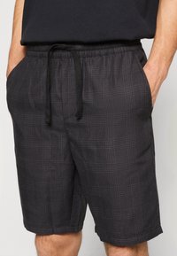 Homme portant un short à carreaux gris foncé avec cordon de serrage, taille élastique et poches latérales, les mains dans les poches, associé à un t-shirt noir.