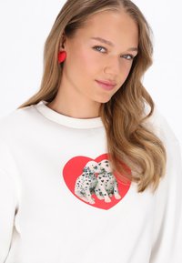 Hanorac alb cu un imprimeu cu doi cățeluși Dalmațieni într-o inimă roșie. Modelul poartă cercei roșii în formă de inimă, cu păr lung și ondulat.