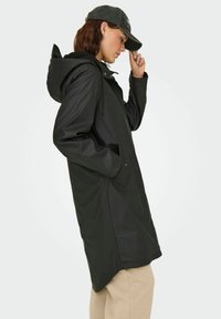 Eine Frau trägt einen schwarzen regenjackenähnlichen Mantel mit Kapuze, hat die Hände in den Taschen und passt eine schwarze Baseballmütze an. Sie steht im Profil vor einem hellen Hintergrund.