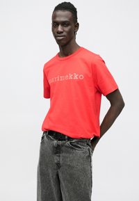 T-shirt de algodão vermelho com o logótipo "marimekko" num tom mais claro, mangas curtas e gola redonda. Combinado com calças de ganga cinzentas.