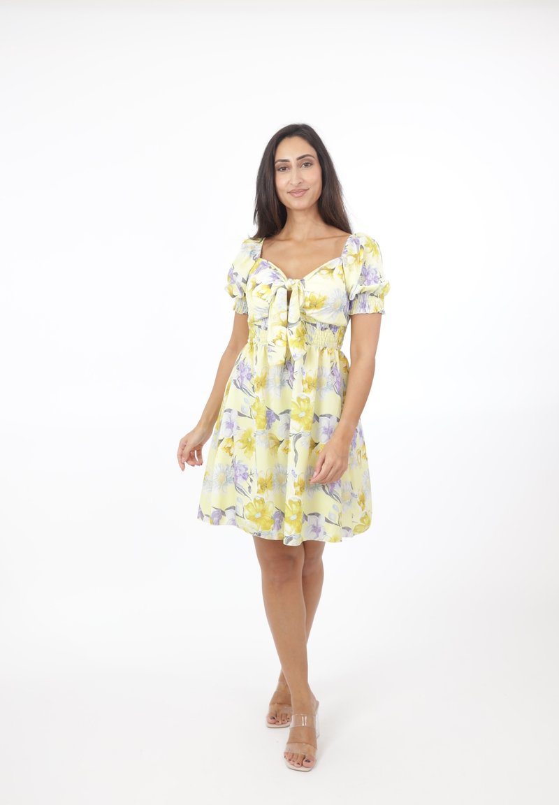 Vestido floral amarillo con detalles morados, con mangas abullonadas, lazo delantero y cintura ajustada, que fluye hacia una falda acampanada.