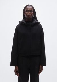 Veste noire courte à capuche avec larges manches, en tissu texturé. Comprend des poches avant et une fermeture dissimulée.