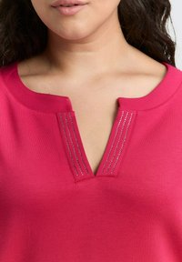 Nahaufnahme einer Frau, die ein leuchtend pinkfarbenes V-Ausschnitt-Oberteil mit kleinen silbernen Verzierungen am Ausschnitt trägt.