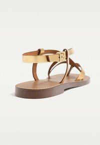 Brune flade sandaler med metalliske guld læderremme og spænde. Har en tekstureret sål og afrundede kanter på en klassisk silhuet.
