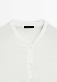 Chemise en lin blanche avec col rond et trois petits boutons, avec une étiquette noire « Massimo Dutti » à l'intérieur du col.