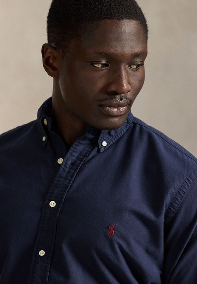 Vyras žiūri į dešinę, dėvi tamsiai mėlyną marškinius su sagomis ir raudoną siuvinėtą logotipą ant krūtinės, neutralaus fono.