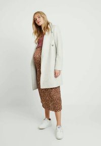 Casaco cinza claro com design de duplo botão, usado sobre uma blusa bordeaux e uma saia midi com bolinhas castanhas, combinado com ténis brancos.