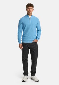 Hellblauer, gerippter Reißverschluss-Pullover mit langen Ärmeln, kombiniert mit dunklen Jeans und grauen Sneakers. Der Pullover hat einen hohen Kragen.