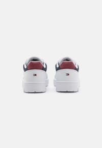 Baskets blanches avec accents navy, un talon rouge et un matériau en cuir texturé. Elles présentent une semelle en caoutchouc blanche et un logo de marque sur le talon.