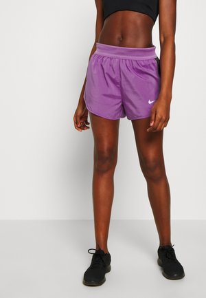 Sportliche Person trägt lila Nike-Shorts, schwarzes Croptop und schwarze Laufschuhe, steht vor weißem Hintergrund.