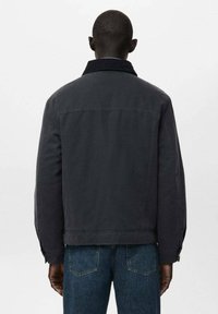 Tmavosivá bunda so štruktúrovaným dizajnom, golierovým výstrihom a zapínanými manžetami, v kombinácii s modrými džínsovými nohavicami. Viditeľné švy.