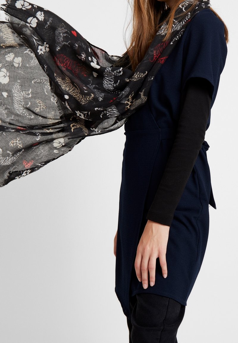 Femme portant une robe bleu marine et un haut à manches longues noir avec une écharpe noire transparente ornée de motifs de tigres, papillons, fleurs et cœurs flottant dans l'air.