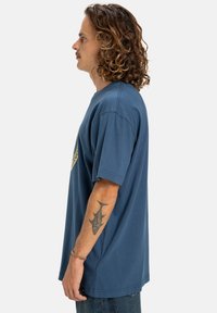 Modré bavlněné tričko s krátkým rukávem, volný střih, kulatý výstřih, s žlutým grafickým designem na hrudi, boční pohled.