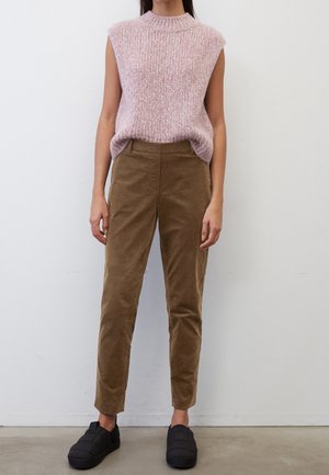 Donna che indossa un top in maglia rosa senza maniche, pantaloni in corduroy marroni e scarpe nere senza lacci, in piedi contro un semplice muro bianco.