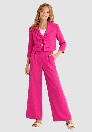 Ragazza giovane in piedi, indossa pantaloni larghi rosa acceso e giacca corta abbinata con fiocco grande, abbinati a una maglietta bianca e sandali.