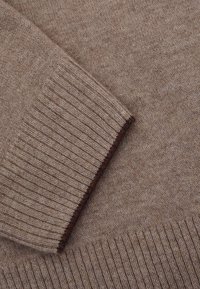 Braunes, geripptes Strickpullover-Detail, das die glatte Textur mit einem dunkelbraunen Akzent am Bündchen zeigt. Das Material erscheint weich und warm.