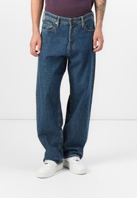 Hombre que lleva jeans oscuros de corte holgado y zapatillas blancas, de pie contra un fondo blanco liso.