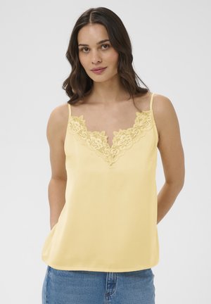 Kvinde iført en lysegul ærmeløs top med blomstret blonderkant på V-halsen, kombineret med blå jeans, stående med hænderne bag ryggen.