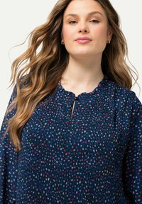 Laivastonsininen paita, jossa on poimuttunut pinta, ominaisuutena aallotettu kaula-auko ja pieni avainreikäyksitys. Koristeltu värikkäillä, hajautetuilla kukkakuoseilla.