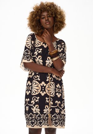 Kobieta o kręconych włosach nosi czarną i beżową sukienkę w kwiatowy wzór, duże kolczyki koła i kilka bransoletek, pozując z ręką przy brodzie.