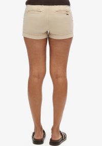 Beige katoenen shorts met opgerolde zomen, voorzien van twee achterzakken en subtiele stiksels. Gecombineerd met donkere slipper sandalen.