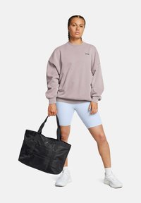 Sweat-shirt oversized violet clair, associé à un short de vélo bleu clair. Sac fourre-tout noir avec poches en maille et logo. Chaussures de sport blanches.