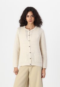 Beige neulejakku, jossa on pyöreä pääntie, napitus, kaksi etutaskua ja teksturoitu kuvio. Suunniteltu pitkille hihoille.