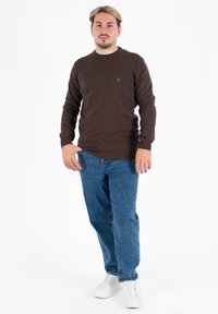 Maglione in maglia marrone con colletto rotondo, abbinato a jeans in denim blu e sneakers bianche. Logo evidente nella zona del petto del maglione.