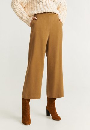 Osoba ubrana w szerokie, camelowe spodnie, beżowe zamszowe botki na obcasie i gruby kremowy sweter z grubym splotem, z rękami w kieszeniach.