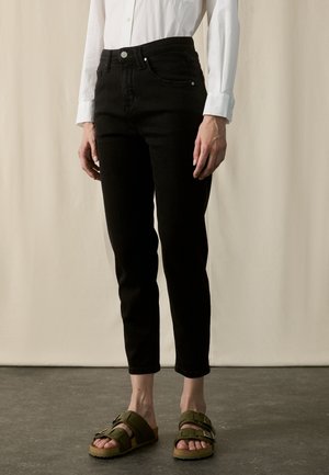 Vrouw draagt zwarte slim-fit jeans, witte blouse met lange mouwen ingestopt en olijfgroene sandalen met gesp, staand op een donkere vloer.