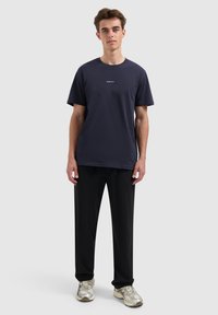 Jonge man staat rechtop, gekleed in een navyblauw "PURE PATH" t-shirt, zwarte broek en beige sneakers tegen een eenvoudige lichte achtergrond.