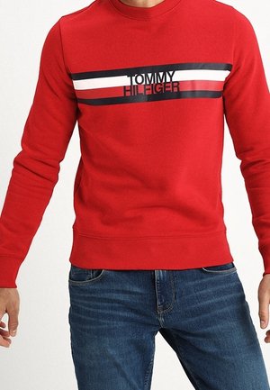 Rotes Sweatshirt mit langen Ärmeln, schwarzen, weißen und roten horizontalen Streifen und dem Schriftzug „Tommy Hilfiger“, kombiniert mit blauen Jeans.