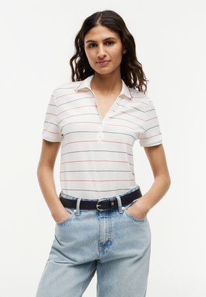 Mladá žena s tmavými vlnitými vlasmi, oblečená v bielom polo tričku s krátkym rukávom a tenkými červenými a čiernymi pruhmi, modré džínsy a čierny opasok, s rukami v vreckách.