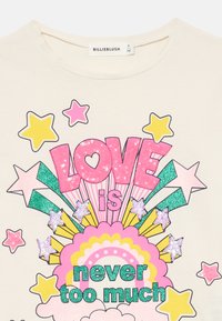 Krém színű ing, amelyet színes csillámos felirat díszít: "A szerelem sosem túl sok", csillagokkal, szivárványokkal és felhőkkel körülvéve, pink, sárga és zöld színekben.