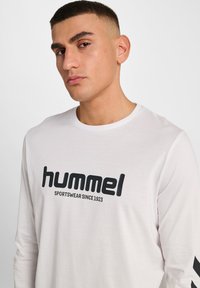 Biała koszula z długim rękawem z dużym czarnym logo "hummel" oraz napisem "SPORTSWEAR SINCE 1923" poniżej. Udekorowana czarnymi paskami na rękawach.