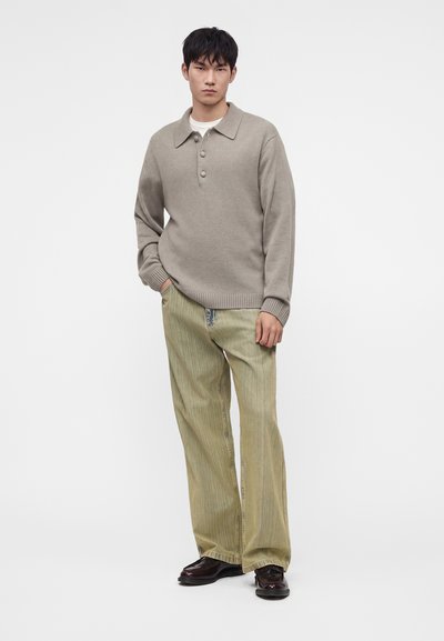 Jeune homme debout portant un pull beige à col boutonné, un pantalon ample vert clair et des chaussures marron foncé sur un fond blanc uni.